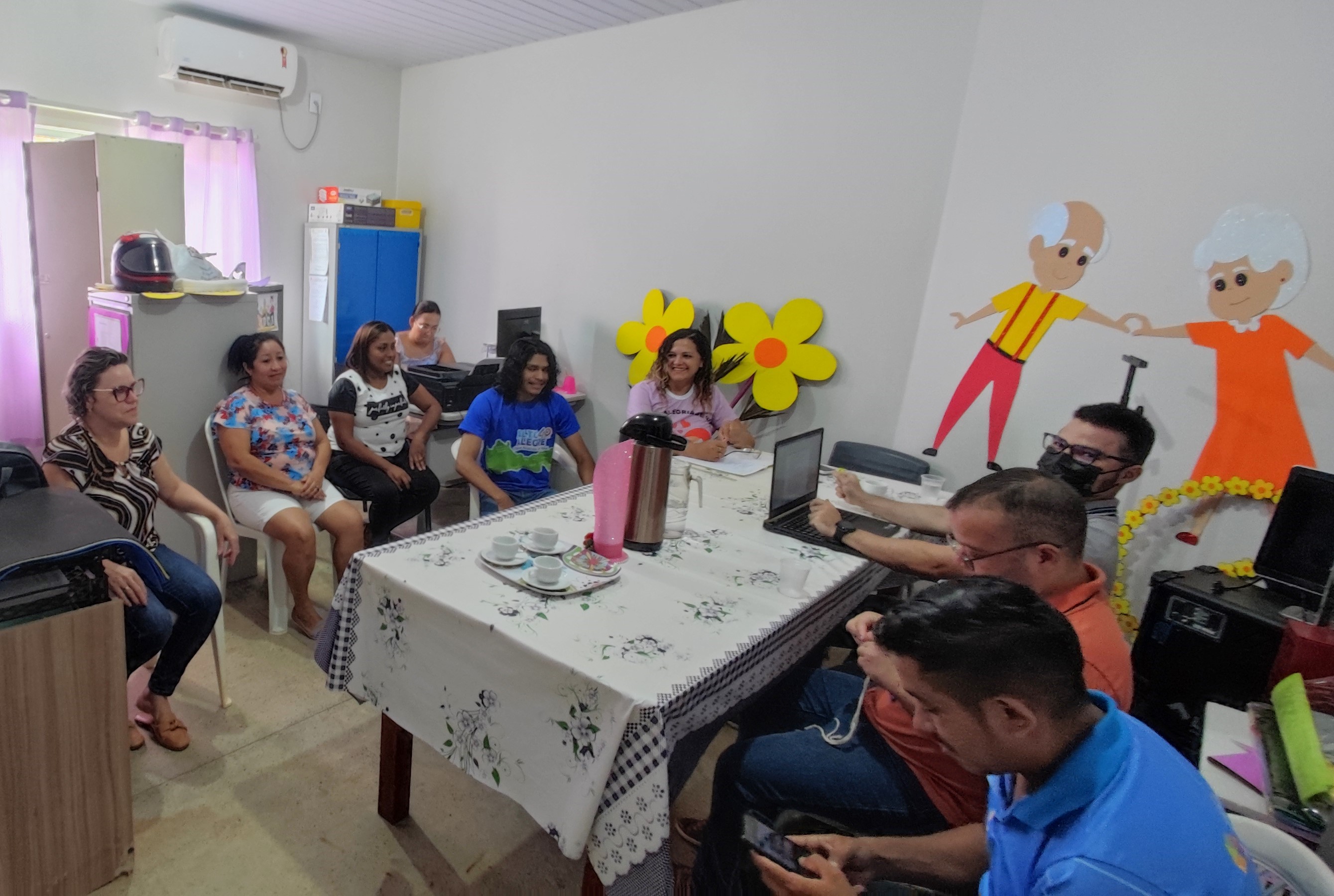 imagem colorida em formato retangular mostra uma sala decorada com cortinas rosas, flores amarelas e desenhos de idosos na parede. No local encontram-se cinco mulheres e quatro homens ao redor de uma mesa, sobre ela há uma garrafa e três xícaras de café.