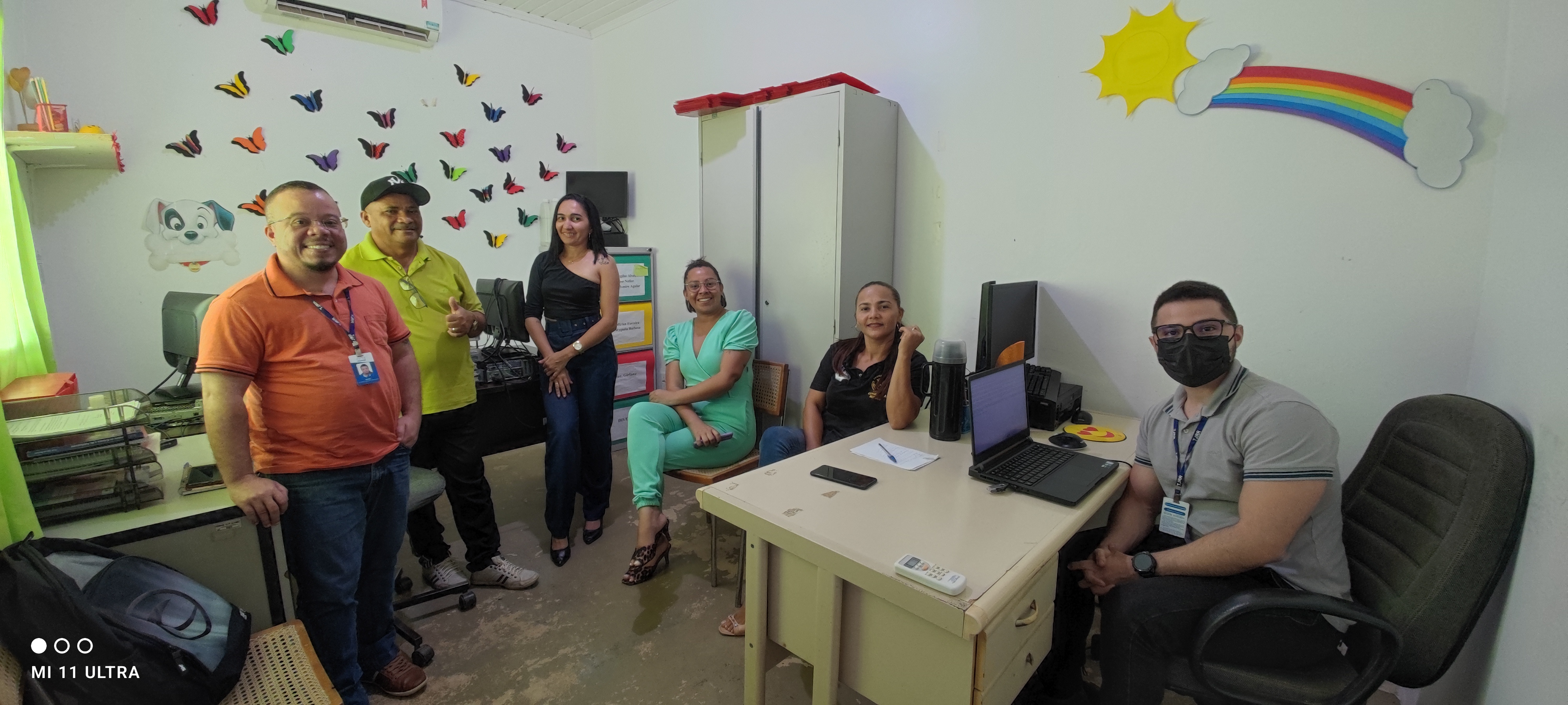 imagem colorida em formato retangular mostra uma sala decorada com borboletas e arco íris. Nela encontram-se três homens e três mulheres, estando alguns sentados e em pé, todos sorrindo e posando para a foto.