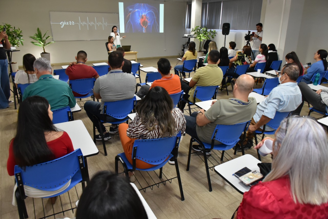 Imagem colorida em formato retangular, mostra uma sala com homens e mulheres sentados em cadeiras escolares na cor azul prestigiando o evento, à frente está a palestrante Glauciene Ferreira de Xavier, mostrando uma uma imagem ilustrativa de um coração e seus vasos sanguíneos.