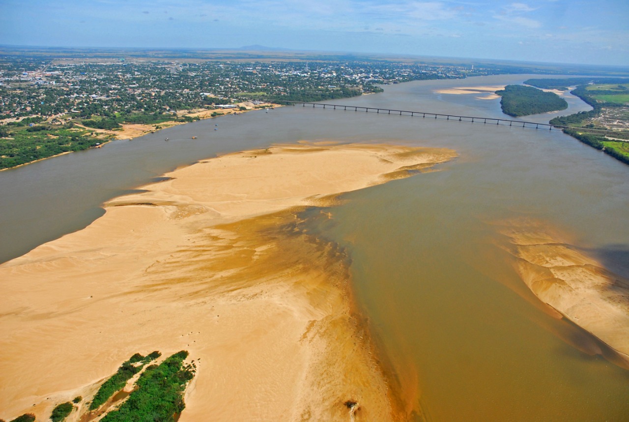 Imagem colorida contém a vista do drone de cima do Rio Branco com bancos de areia e ao fundo da imagem contém a Ponte dos Macuxis.