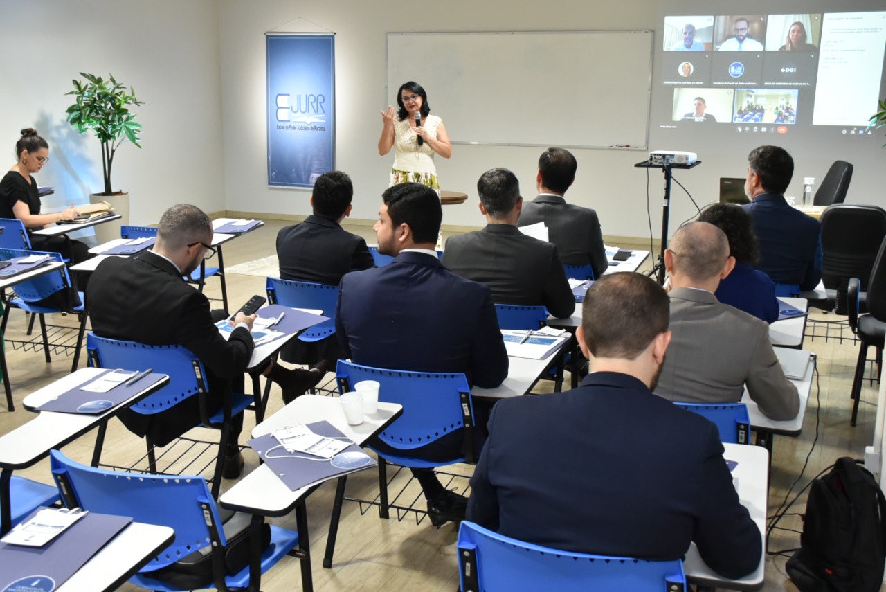 Imagem colorida mostra os magistrados e magistradas sentados em carteiras de tons azul e branco dentro de uma sala com um quadro branco ao lado do quadro há uma projeção de uma sala virtual com oito participantes, na frente do quadro está a mediadora do evento em pé com o microfone na mão.