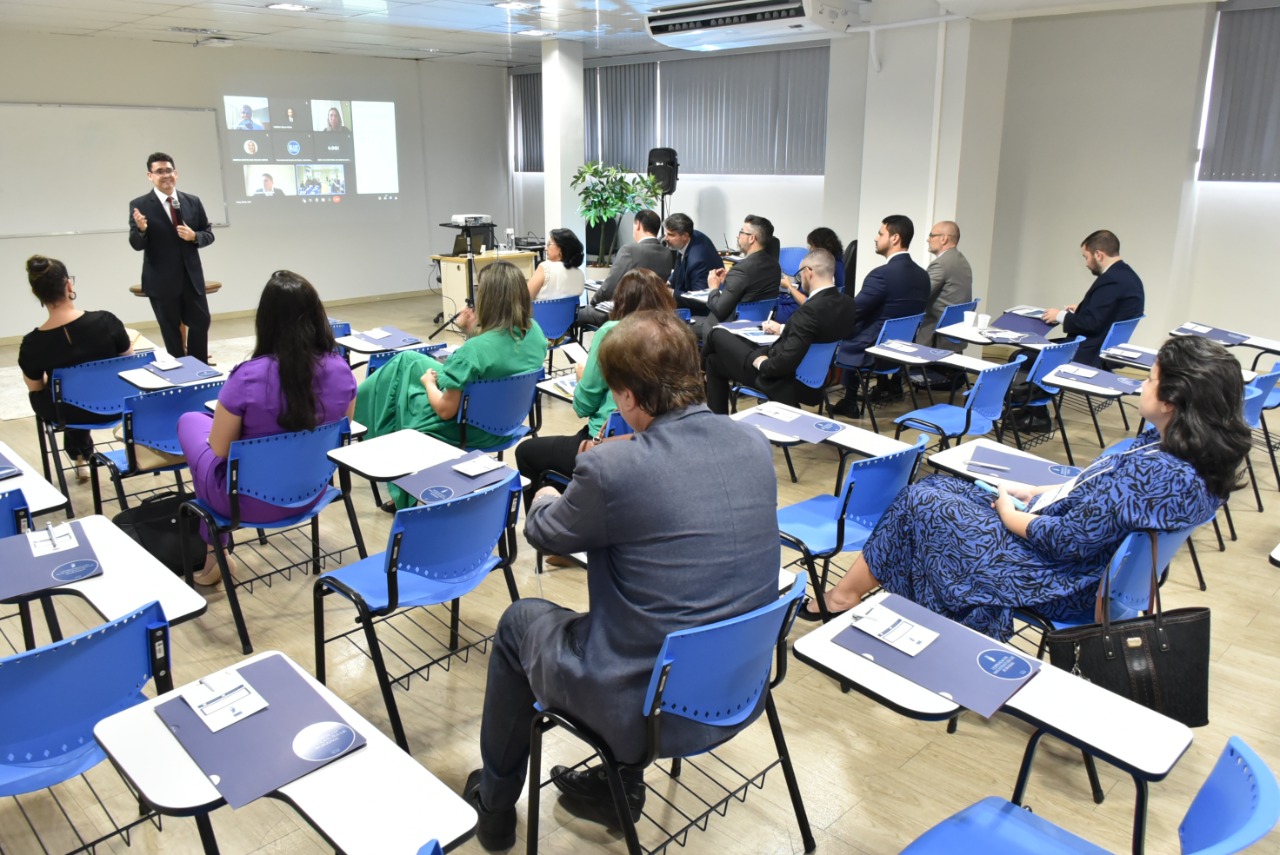 Imagem colorida mostra os magistrados e magistradas sentados em carteiras de tons azul e branco dentro de uma sala com um quadro branco ao lado do quadro há uma projeção de uma sala virtual com oito participantes, na frente do quadro está o mediador do evento em pé com o microfone na mão.