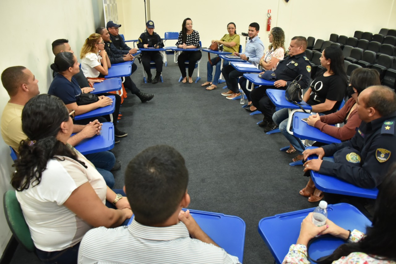 Imagem colorida mostra um auditório, onde o grupo de guardas civis municipais que integram a Patrulha Maria da Penha estão sentados em carteiras escolares formando um círculo, a juíza Suellen Alves está sentada entre o grupo vestindo uma camisa preta com bolinhas brancas, a mesma está facilitando o evento.