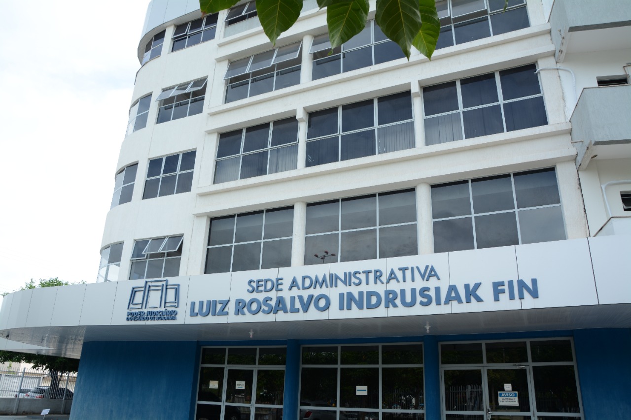 Imagem colorida mostra a fachada do Prédio da Sede Administrativa Luiz Rosalvo Indrusiak Fin, o edifício está identificado com o nome do prédio pintado de azul escuro, possui 4 andares com janelas de vidros  e um térreo.