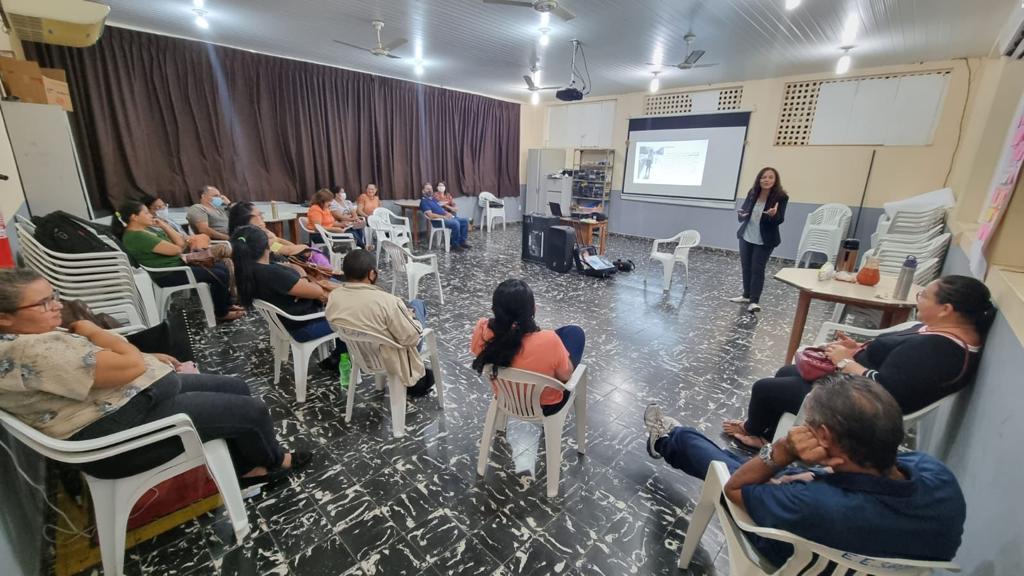 Imagem mostra os gestores e professores sentados em  uma sala com cadeiras brancas  coladas em formação de círculo, à frente está  a coordenadora e psicóloga do programa, facilitando o evento.