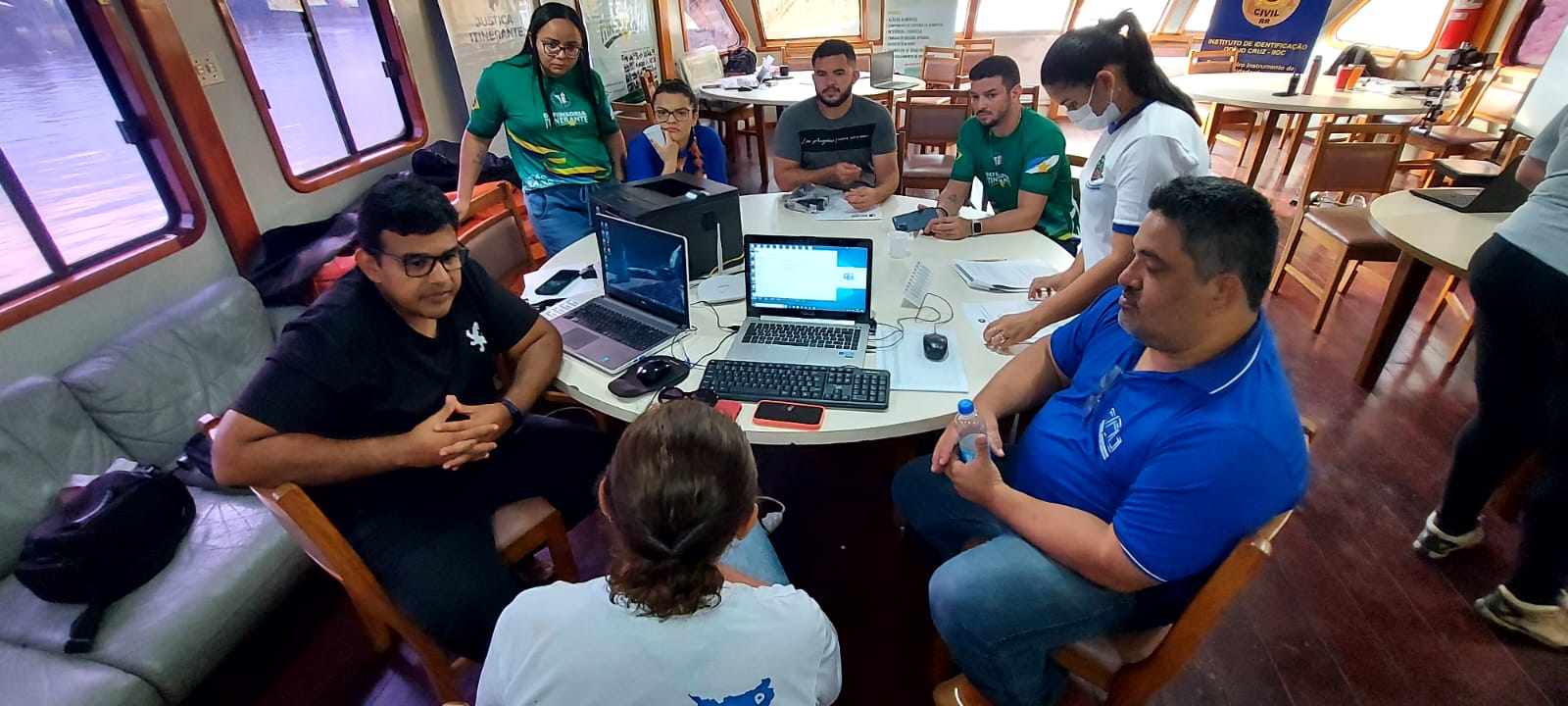 Imagem colorida mostra uma sala com 8 pessoas reunidos ao redor de uma mesa redonda, sob a mesa estão documentos, celulares e notebooks.