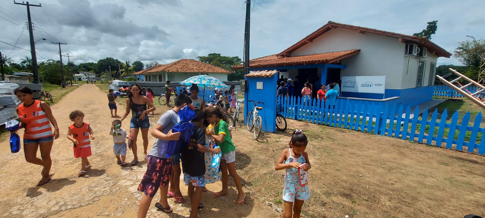 Imagem colorida mostra a fachada do prédio da Justiça Cidadã da comunidade Santa Maria do Boiaçu, o edifício pintado nas cores branco e azul escuro está identificado com o nome e com a logo do Tribunal de Justiça de Roraima e possui ao redor cercas de madeira no tom azul escuro. Na frente há crianças desembrulhando  presentes, além de homens e mulheres saindo do prédio.  