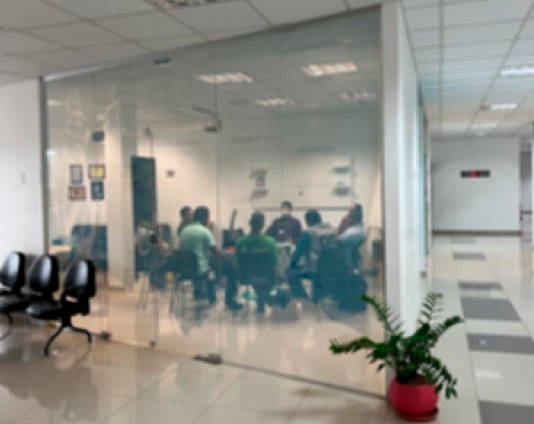 Imagem colorida embaçada mostra o encontro restaurativo para autores de violência doméstica em uma sala com paredes de vidro transparente. Na foto estão 9 homens, sendo um deles o mediador do evento.