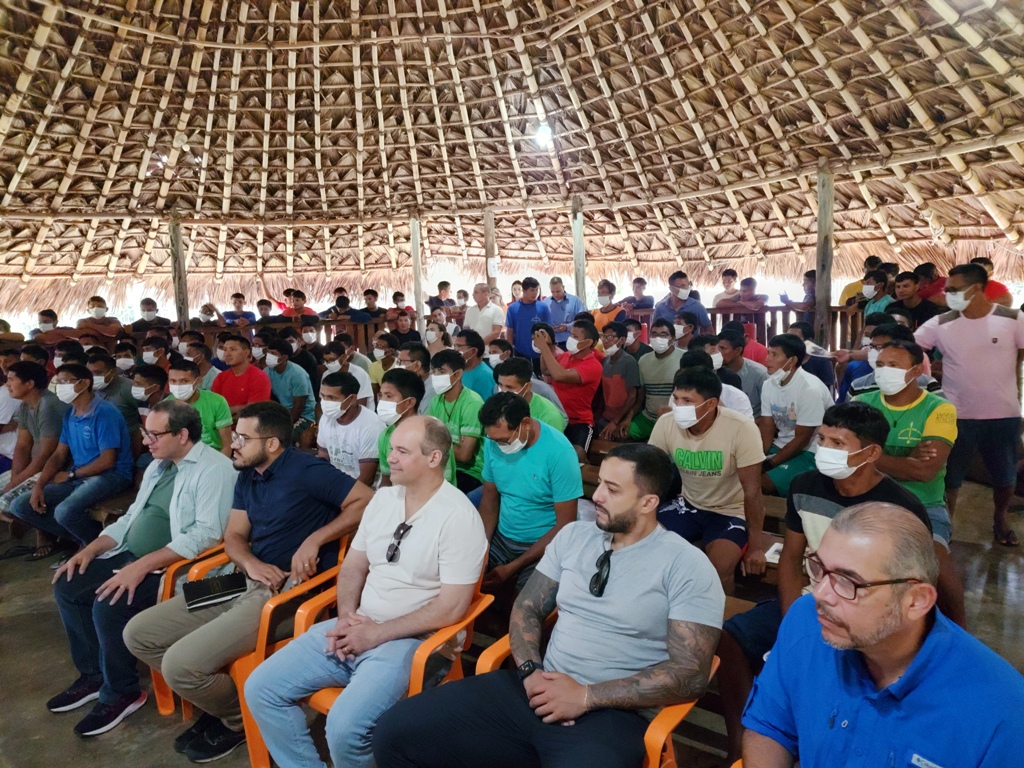  Imagem colorida mostra um malocão com homens e mulheres da comunidade Indigena Waimiri Atroari, juntamente com magistrados, magistradas, servidores, servidoras, sentados prestigiando o evento.