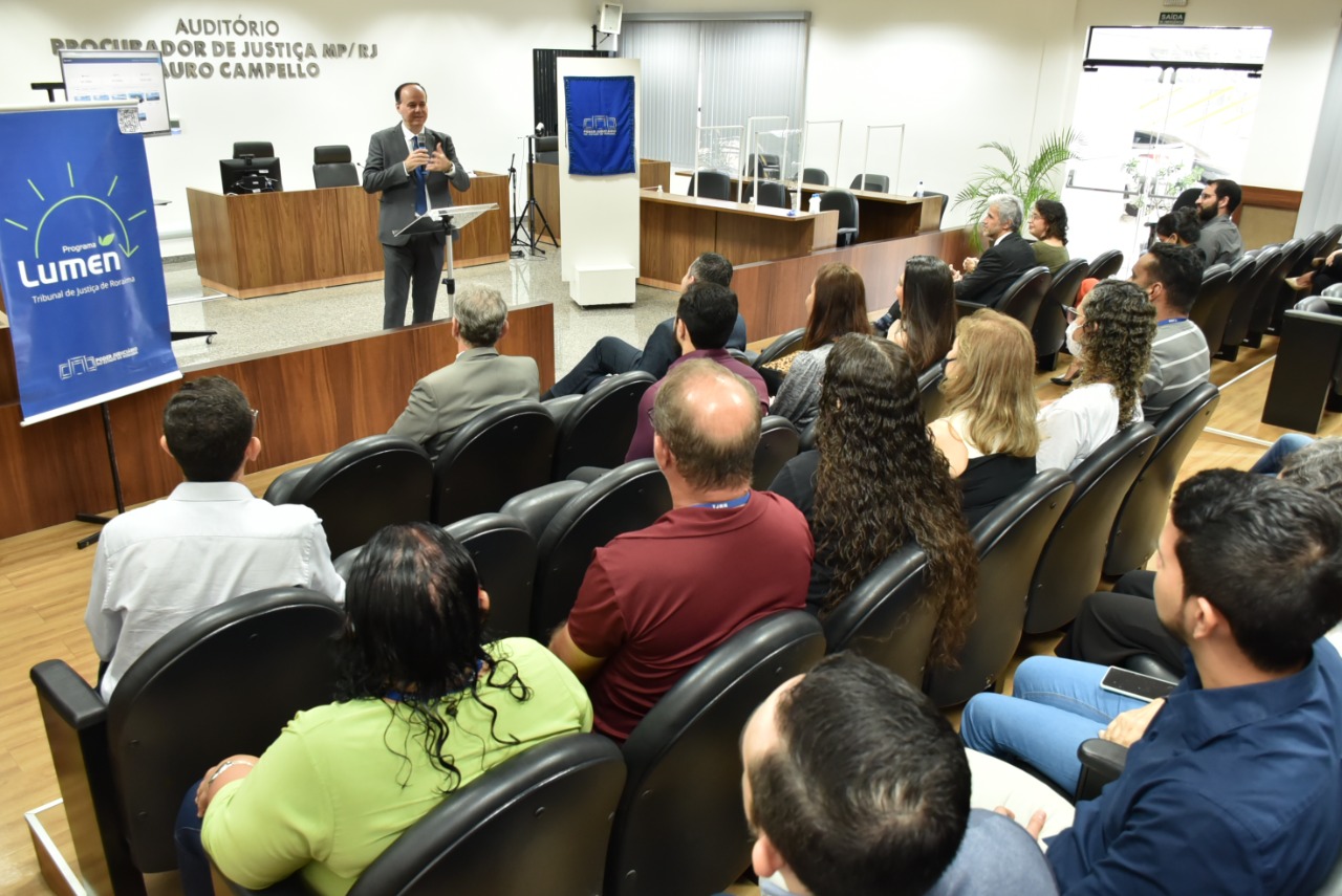 Imagem colorida mostra o presidente TJRR, desembargador Cristóvão Suter, discursando em um auditório para um grupo de homens e mulheres, a foto mostra também, dois banners azuis, um com a logo do TJRR e outra com a logo do projeto lumen, ambas estão posicionadas nas laterais à frente da plateia.