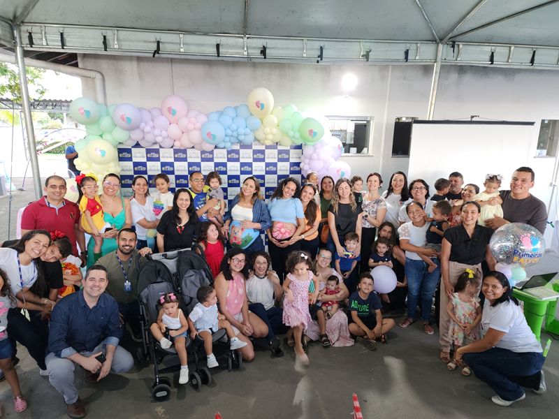 Imagem 1- Imagem colorida em formato retangular, mostra pais, mães, mães gestantes e crianças entre 0 a 3 anos de idade, sorridentes. posando para a foto em frente a um painel decorado com balões coloridos