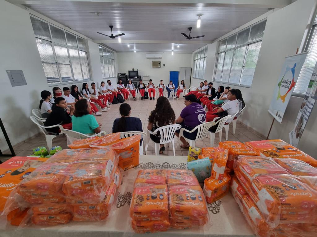  Imagem colorida mostra uma sala branca com janelas de vidros, no centro da foto há alunos, alunas e servidoras da  Escola Estadual Oswaldo Cruz, sentados em cadeiras brancas formando um círculo, na parte inferior da foto há uma mesa com diversos pacotes de fraldas. 