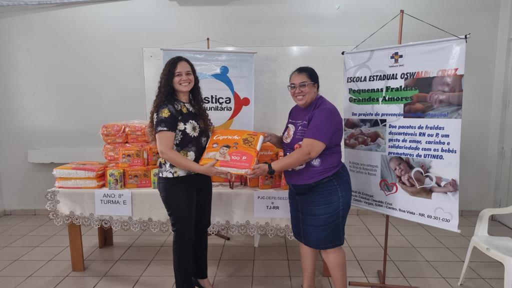 Imagem colorida em formato retangular, mostra duas servidoras da Escola Estadual Oswaldo Cruz segurando um pacote de fraldas, atrás encontra-se uma mesa com vários pacotes de fraldas e dois cartazes de divulgação.