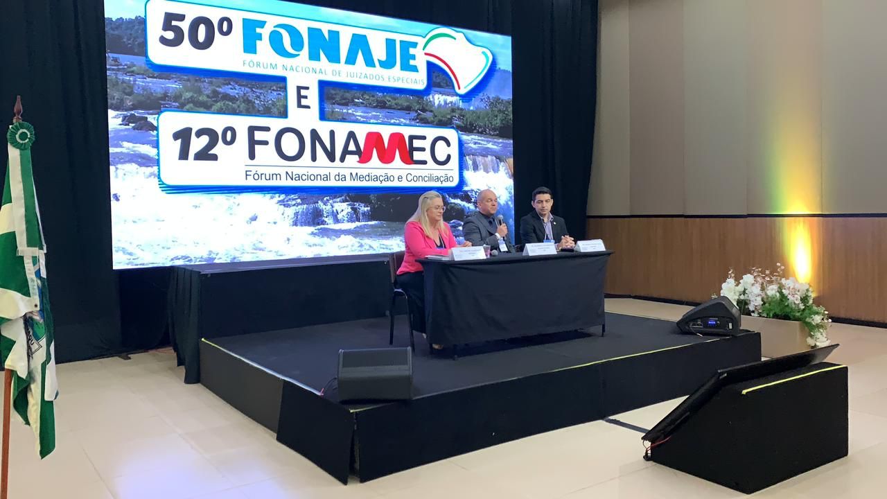 Imagem colorida mostra mesa de autoridades composta por dois homens e uma mulher, atrás exibida em um telão está a imagem ilustrativa do foz do iguacú e no centro em destaque  o texto “50° FONAJE E 12° FONAMEC”