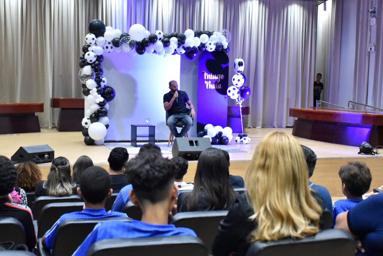 Imagem colorida em formato retangular mostra o auditório do Fórum Cível Advogado Sobral Pinto com um público de homens, mulheres, jovens, adolescentes e crianças durante a palestra “Ouro Norte”, ministrado pelo jogador do Flamengo, Thiago Maia, o mesmo estar a frente da plateia sentado , atrás há um painel decorado com balões nas cores preto e branco e eles alguns balões simulam bolas de futebol.