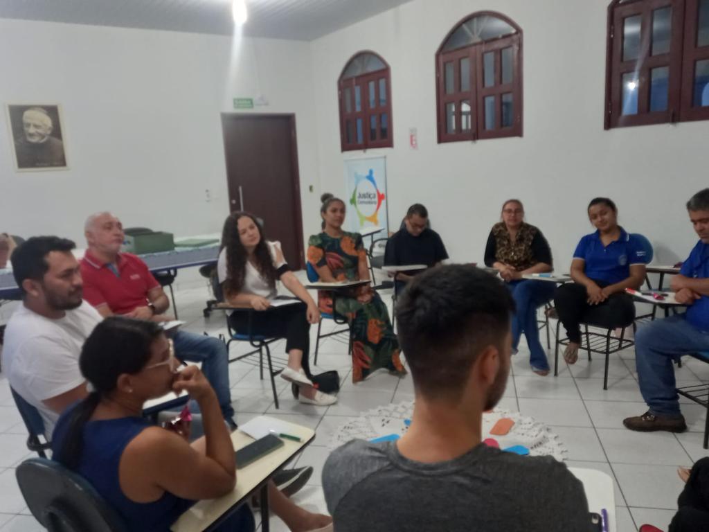 Imagem colorida em formato retangular mostra um sala com paredes brancas e janelas e portas pintadas de vermelho escuro e um grupo composto por homens e mulheres sentados em cadeiras formando um círculo. 