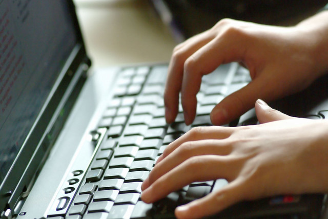 Imagem ilustrativa colorida mostra mãos femininas realizando a ação de digitar em um teclado de um notebook.