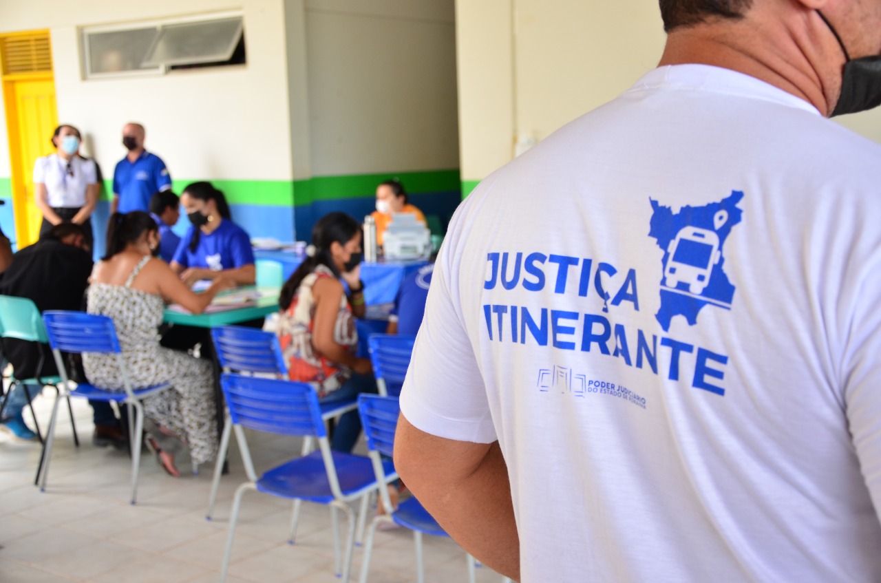 Imagem colorida em formato retangular com o fundo desfocado mostra um servidor de costas com uma camisa branca estampada com o logotipo da vara da justiça itinerante.