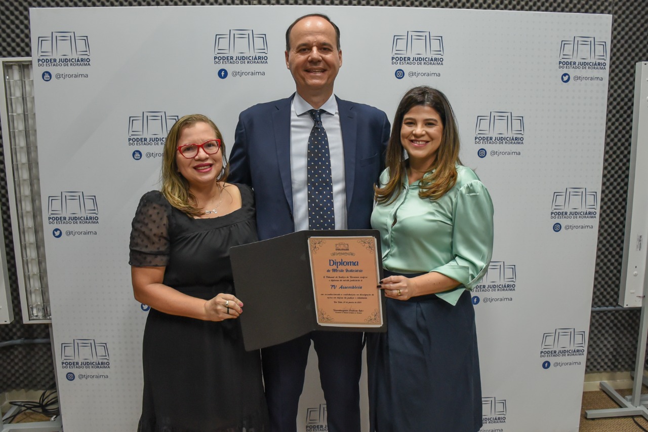 Imagem colorida em formato retangular mostra o presidente do TJRR, desembargador Cristóvão Suter ao lado da superintendente de Comunicação da TV Assembléia, Sônia Lúcia Nunes Pinto e a jornalista Camilla Dall´Agnol, posando em pé para foto com o diploma de “Mérito Judiciário” em suas mãos.