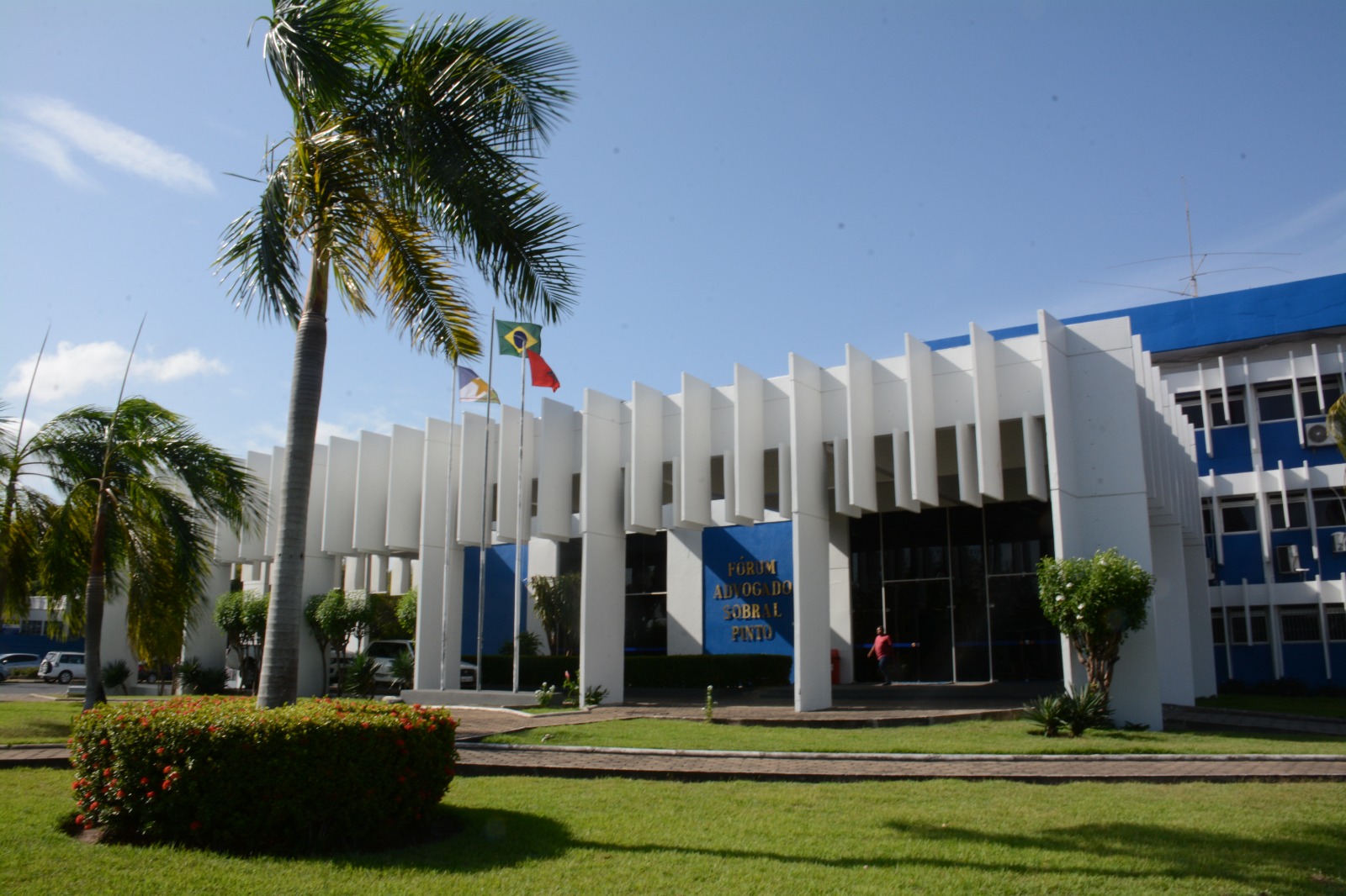  Imagem colorida em formato retangular mostra a fachada arborizada do Fórum Advogado Sobral Pinto, o prédio possui três andares e  está pintado nos tons branco e azul.