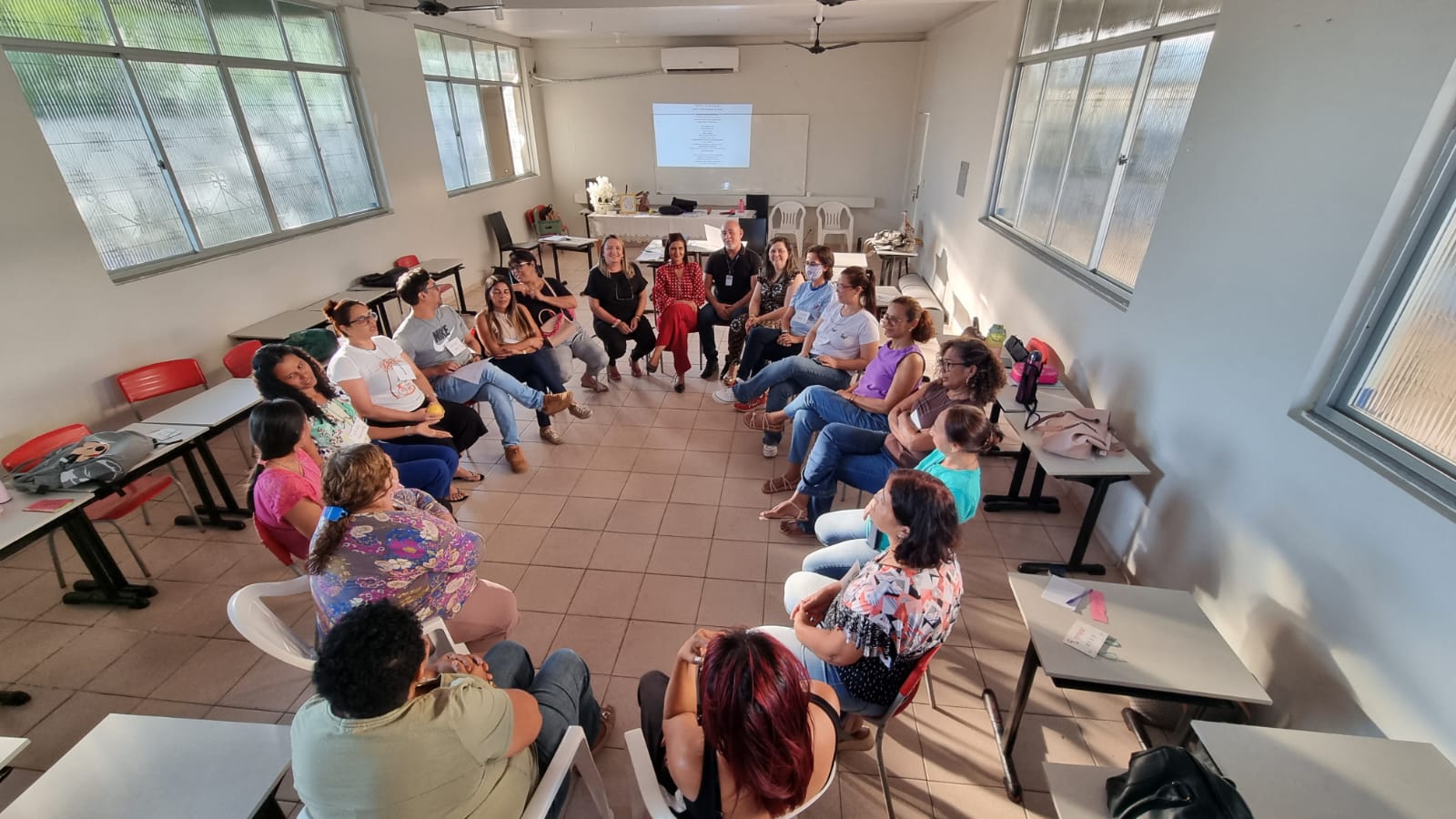 Imagem colorida em formato retangular mostra uma sala branca com cadeiras escolares ao redor. No centro foram colocadas cadeiras brancas que formam um círculo, ocupadas por homens e mulheres, colaboradores da escola Estadual Oswaldo Cruz.