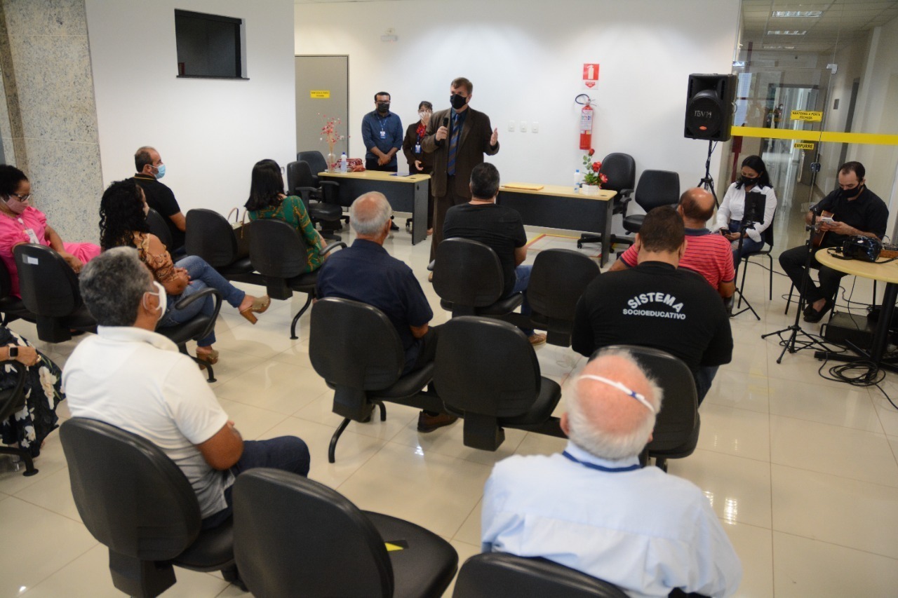 Imagem colorida em formato retangular, mostra uma sala com cadeiras na cor preta ocupadas por homens e mulheres prestigiando um evento, à frente facilitando o evento está o juiz Alexandre Magno, titular da Vepema.
