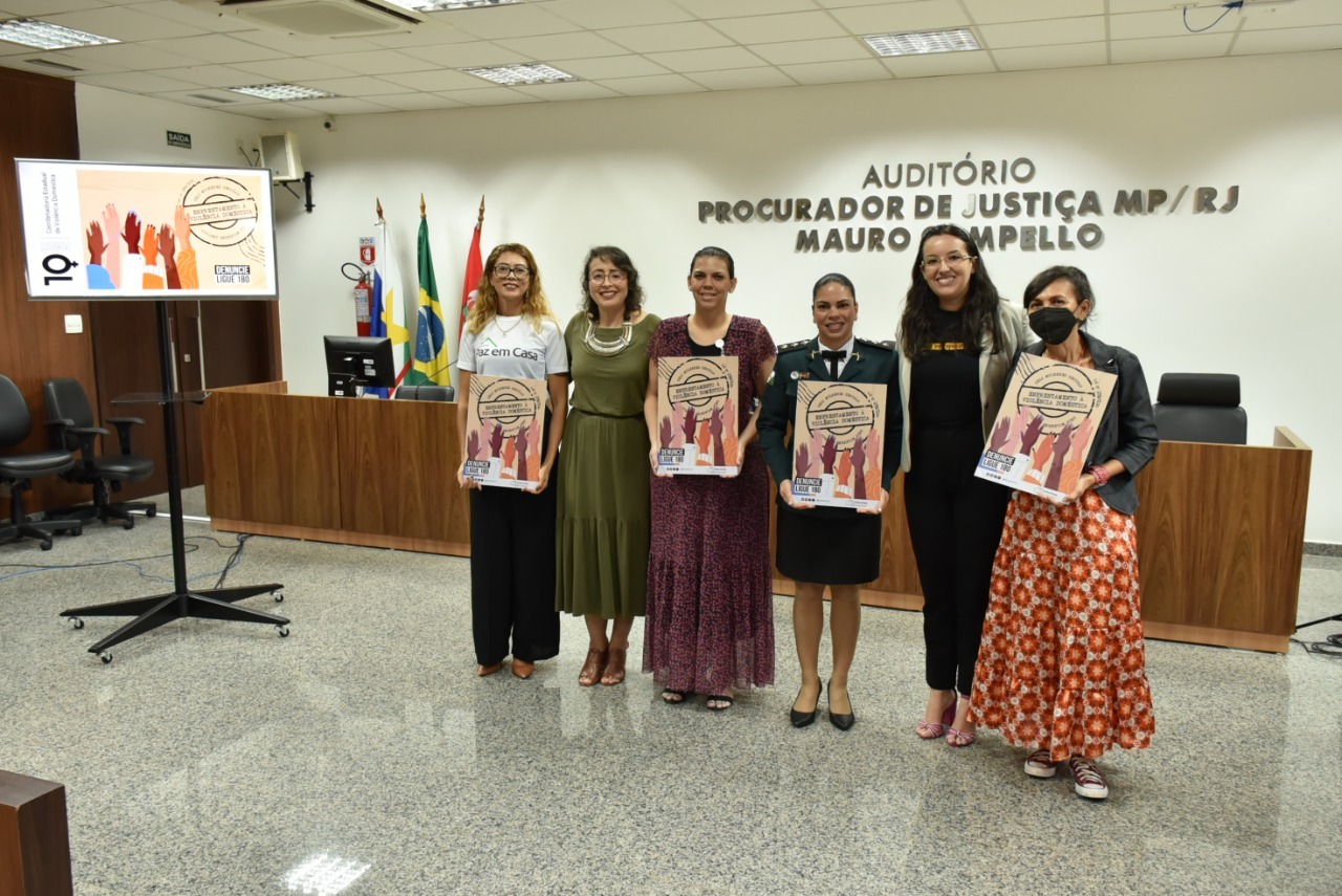 Imagem colorida em formato retangular mostra a coordenadora Estadual da mulher em situação de violência doméstica e familiar do TJRR, Juiza Suelen Alves  a diretora do Fórum Criminal Lana Leitão, a professora Mara Maia, a criadora do aplicativo “Solte a Voz”, Nanníbia Oliveira Cabral, a coordenadora do Programa Observatório da Violência contra a Mulher em RR da Universidade Federal de Roraima, Luziene Corrêa Parnaíba e a major Dyanna Vieira de Oliveira da Polícia Militar de Roraima, posando para a foto, as ganhadoras  estão segurando o selo de reconhecimento mulheres seguras.