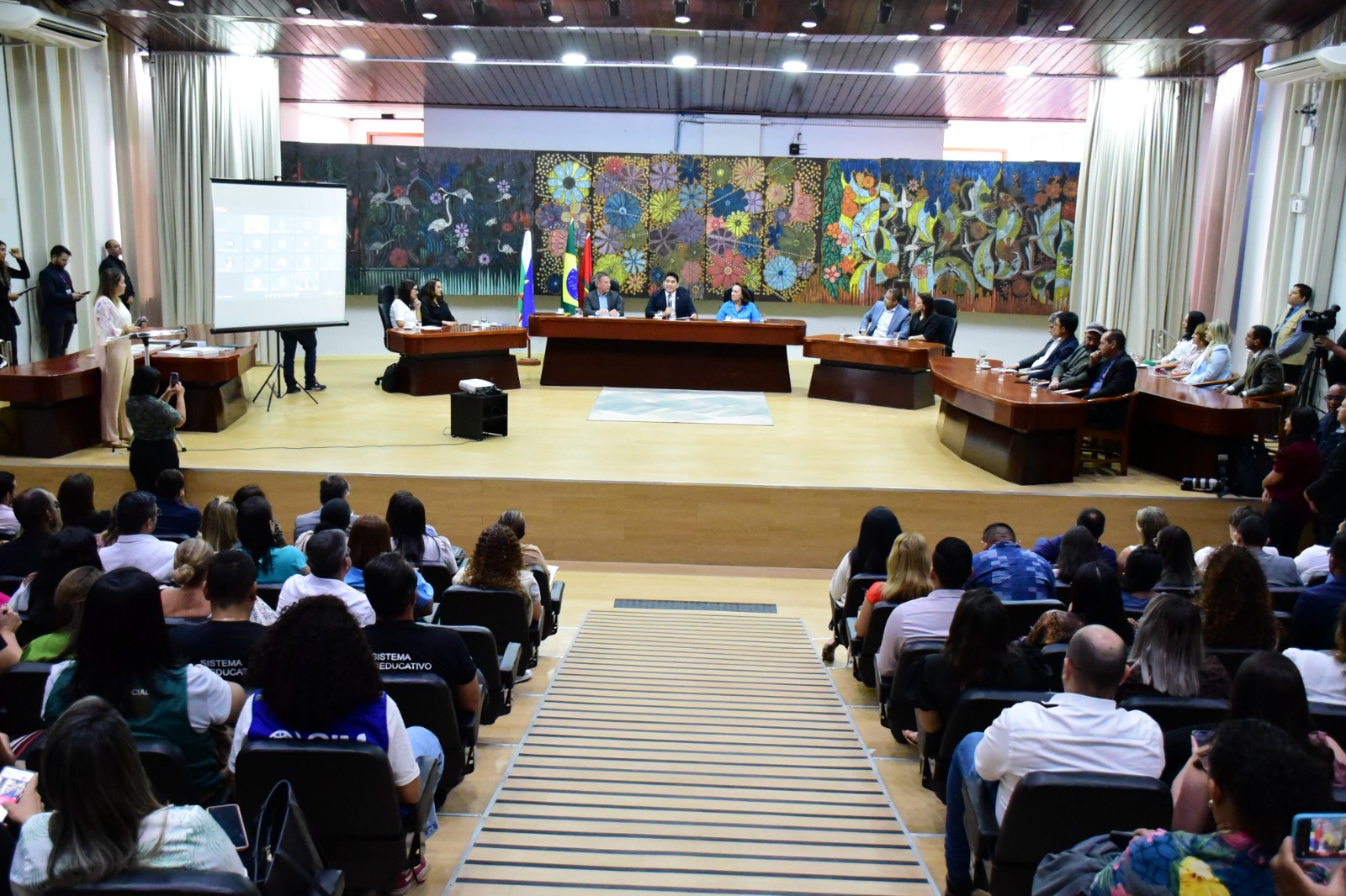  Imagem colorida mostra o auditório do Fórum cível ocupado por servidora e servidores do Poder Judiciário de Roraima, na frente na mesa de autoridades estão os 14 representantes de instituições públicas. 