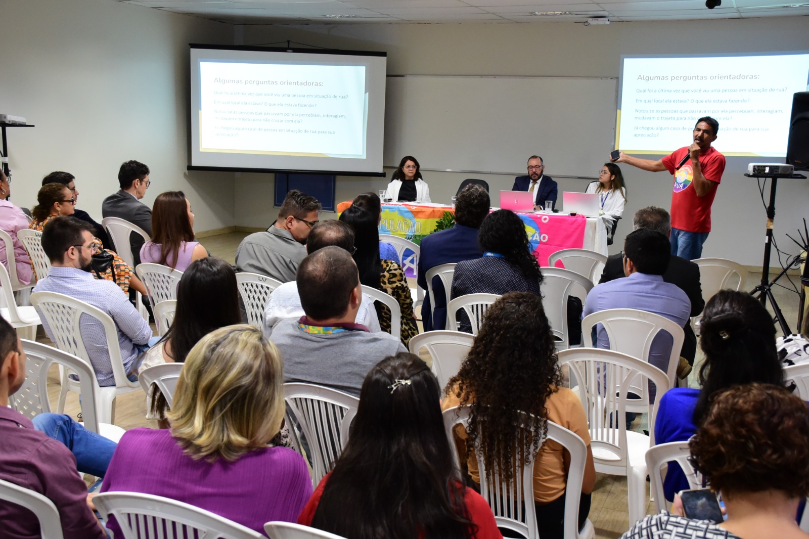 Imagem colorida mostra uma sala com cadeiras brancas ocupadas por homens e mulheres. Ao fundo, mesas cobertas com a bandeira do Movimento Nacional População de Rua (MNPR). No canto superior esquerdo em pé, com o microfone na mão está o membro do Movimento Nacional População de Rua (MNPR), Vanilson Torres.