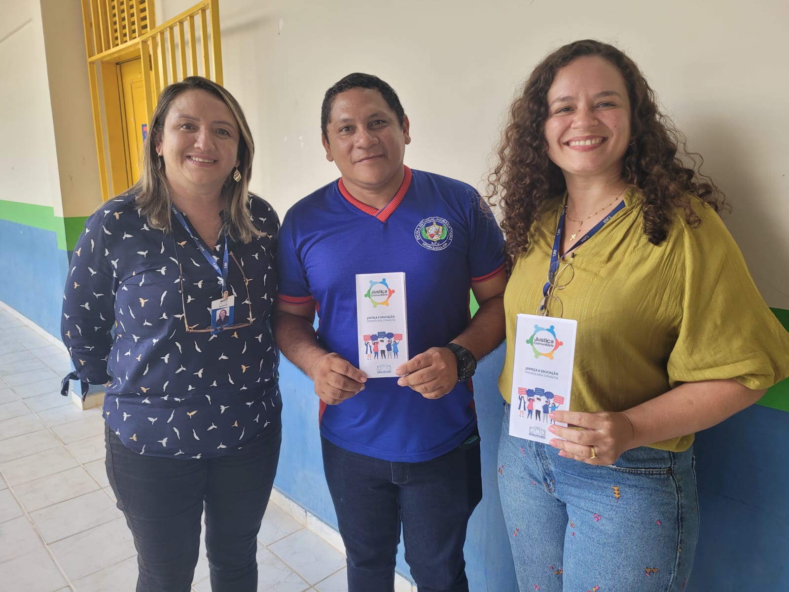 Imagem colorida mostra a coordenadora do Programa Justiça Comunitária, Marcelle Wottrich e a mediadora Dilma Roselli Coutinho, posando para foto ao lado de um educador .