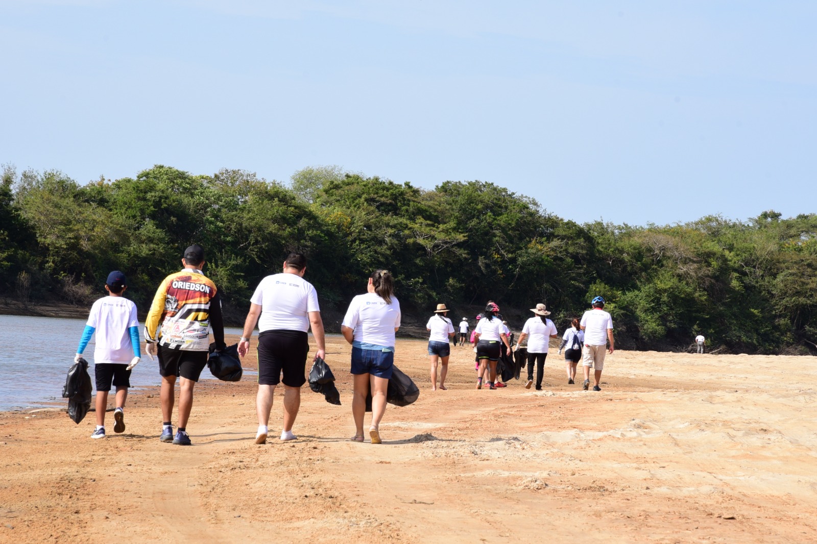 Imagem colorida mostra um grupo de pessoas de costas, vestindo camisas brancas e segurando um saco preto na mão, caminhando pela praia do Caçari.