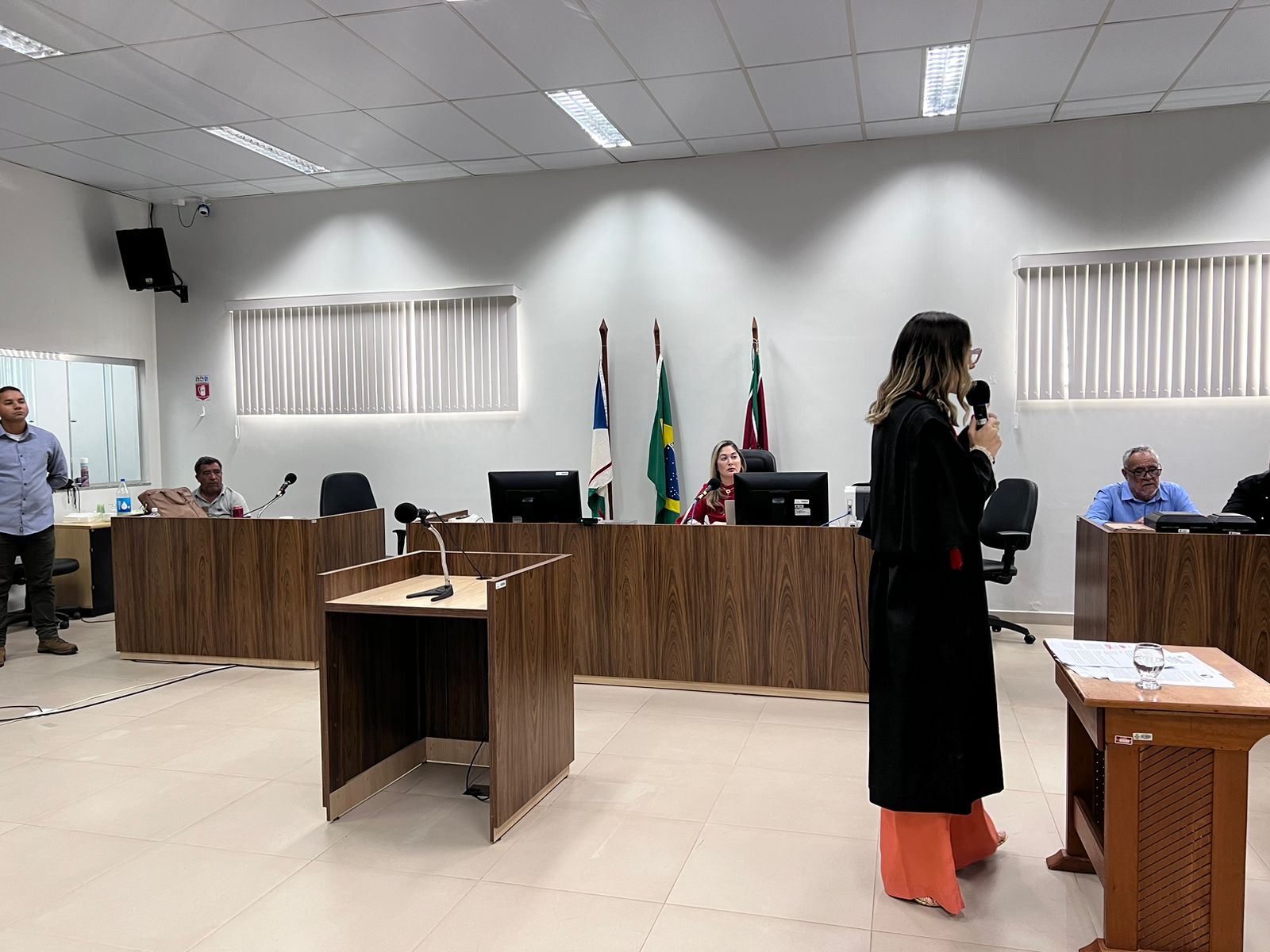 Imagem colorida mostra uma sessão de julgamento do Tribunal do Júri na Comarca do Município de São Luiz. No lado esquerdo da foto está uma magistrada usando toga , posicionada lateralmente com o microfone na mão.