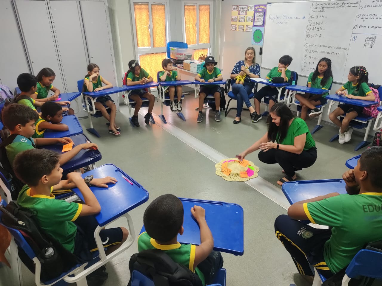 Imagem colorida mostra uma sala de aula com carteiras escolares azuis, posicionadas formando um círculo, nelas estão alunos e alunas fardados com camisas verdes com golas amarelas. No centro, uma servidora coloca um post it sobre um tapete de crochê redondo.