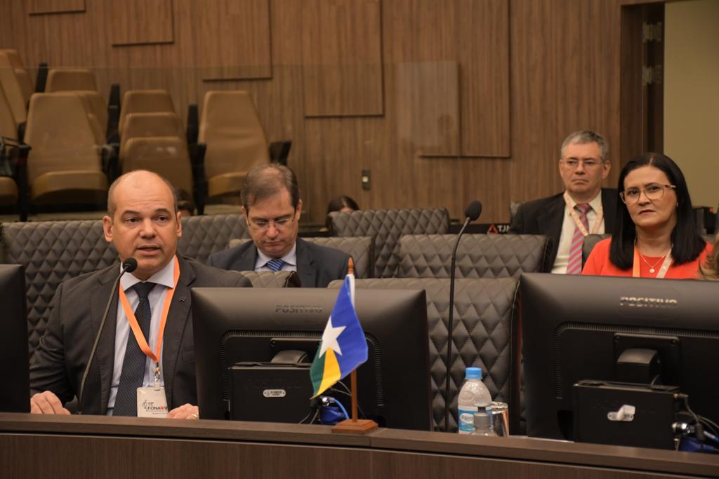 Imagem colorida mostra o juiz Eduardo Álvares Carvalho, coordenador do Centro Judiciário de Solução de Conflitos e Cidadania do Tribunal de Justiça de Roraima (CEJUSC/TJRR), durante debate. A frente está o microfone e uma pequena bandeira do estado, ao fundo estão dois homens e uma mulher sentados em cadeiras pretas.
