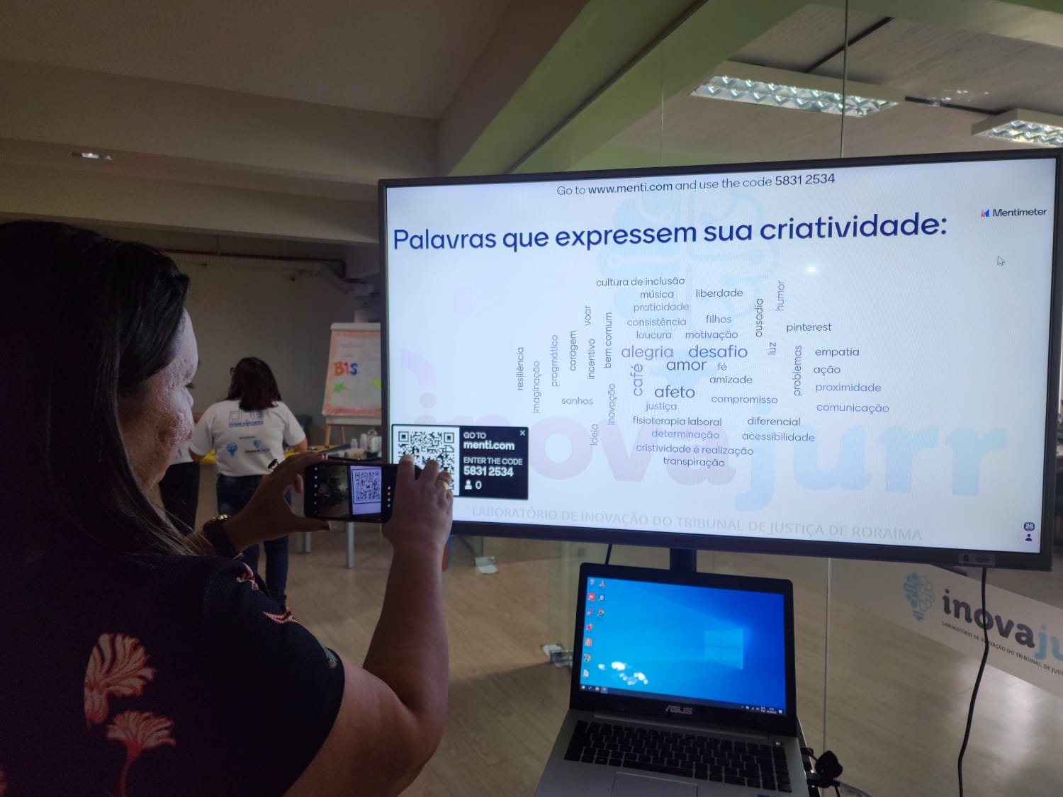 Imagem colorida mostra uma mulher com o celular na mão escaneando um qr code em uma tela de tv que exibe diversas palavras, em destaque está a frase “ Palavras que expressem sua criatividade”
