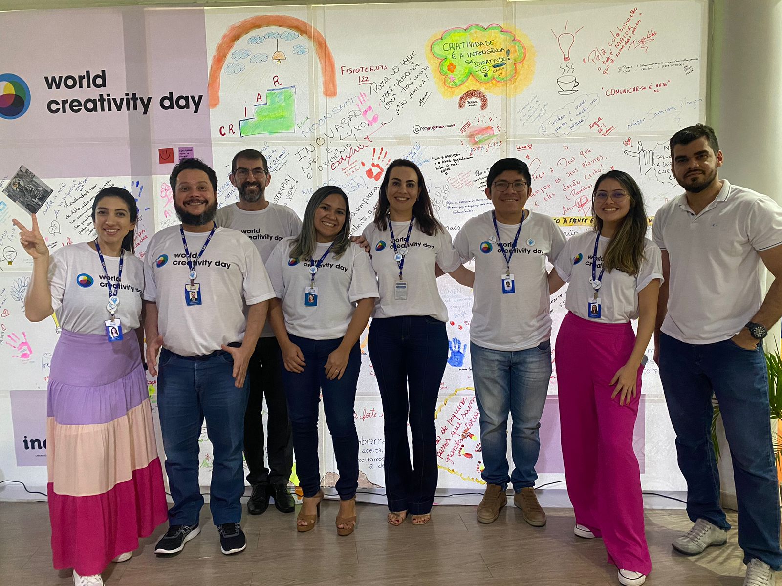 Imagem colorida mostra 8 servidores, em pé, posando para foto à frente de uma painel decorado com frases e desenhos coloridos