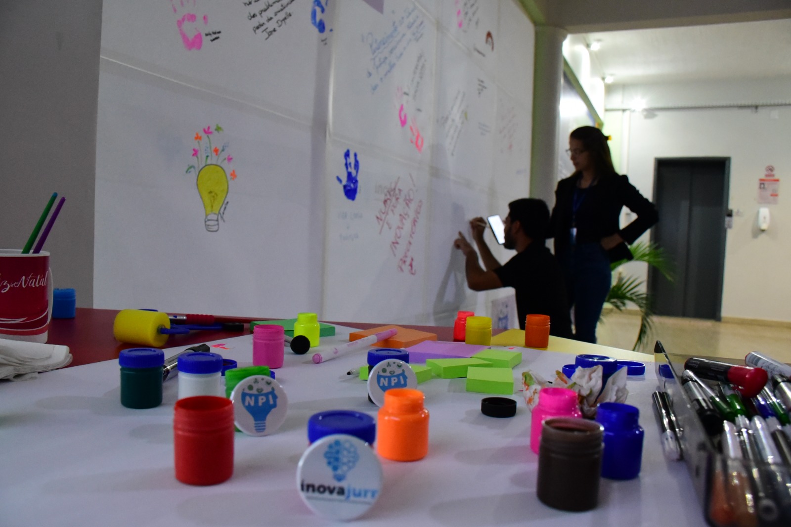Imagem colorida mostra uma mesa com diversos potes de tinta guache, post-its e canetas coloridas. Ao fundo há um homem agachado escrevendo em uma parede com cartolinas brancas decoradas com mãos e frases, ao seu lado, em pé, está uma mulher observando a ação.