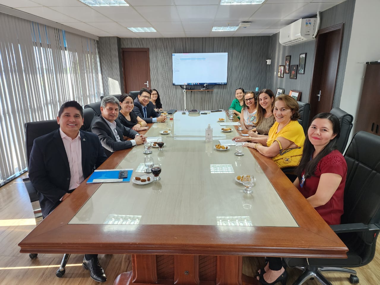 oto colorida mostra o presidente do TJRR, desembargador Jésus Nascimento, reunido na mesa de reunião da sala da presidência com os juízes titulares das Varas da Infância e Juventude, Parima Dias Veras e Marcelo Oliveira, a juíza titular da Vara da Justiça Itinerante, Graciete Sotto Mayor, a conselheira do TCE, Cilene Salomão e a defensora pública Emira Salomão. Além da secretária e a assessora executiva da Secretaria de Gestão de Magistrados, Fernanda Catanhede e Franciza Veríssimo, a assessora Ana Moreira e a assessora executiva da Secretaria de Infraestrutura e Logística do Poder Judiciário de Roraima, Claudete Silva. Todos estão posando e sorrindo para a foto. Na sala também estão objetos como televisão e quadros artísticos. Abaixo está o texto “PROJETOS SOCIAIS. Presidente do TJRR reúne TCE, DPE e magistrados para debater revitalização de prédio público