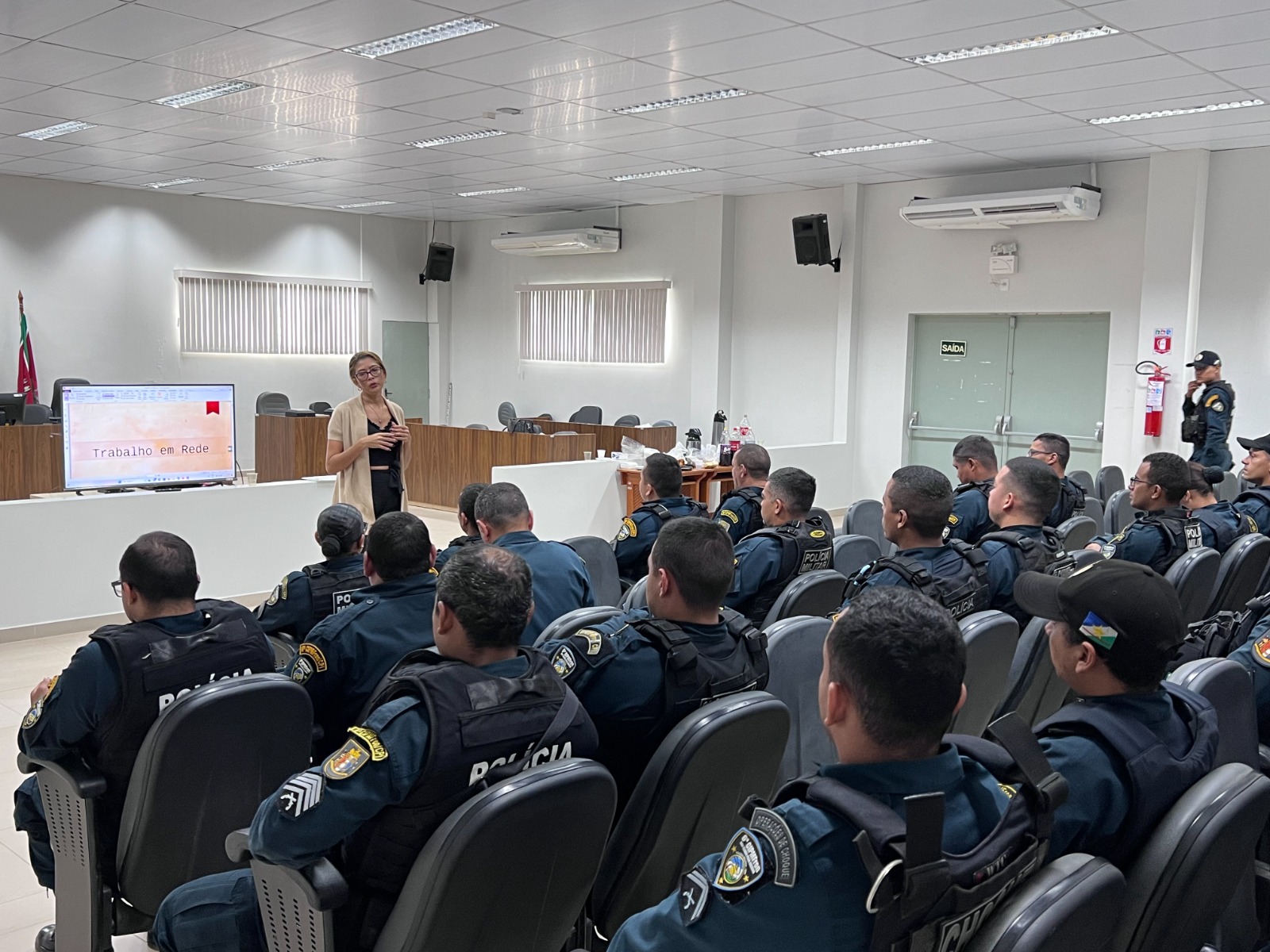 FOTO COLORIDA DE UM AUDITORIO COM POLICIAIS SENTADOS ENQUANTO UMA SERVIDORA MINISTRA A CAPACITAÇÃO