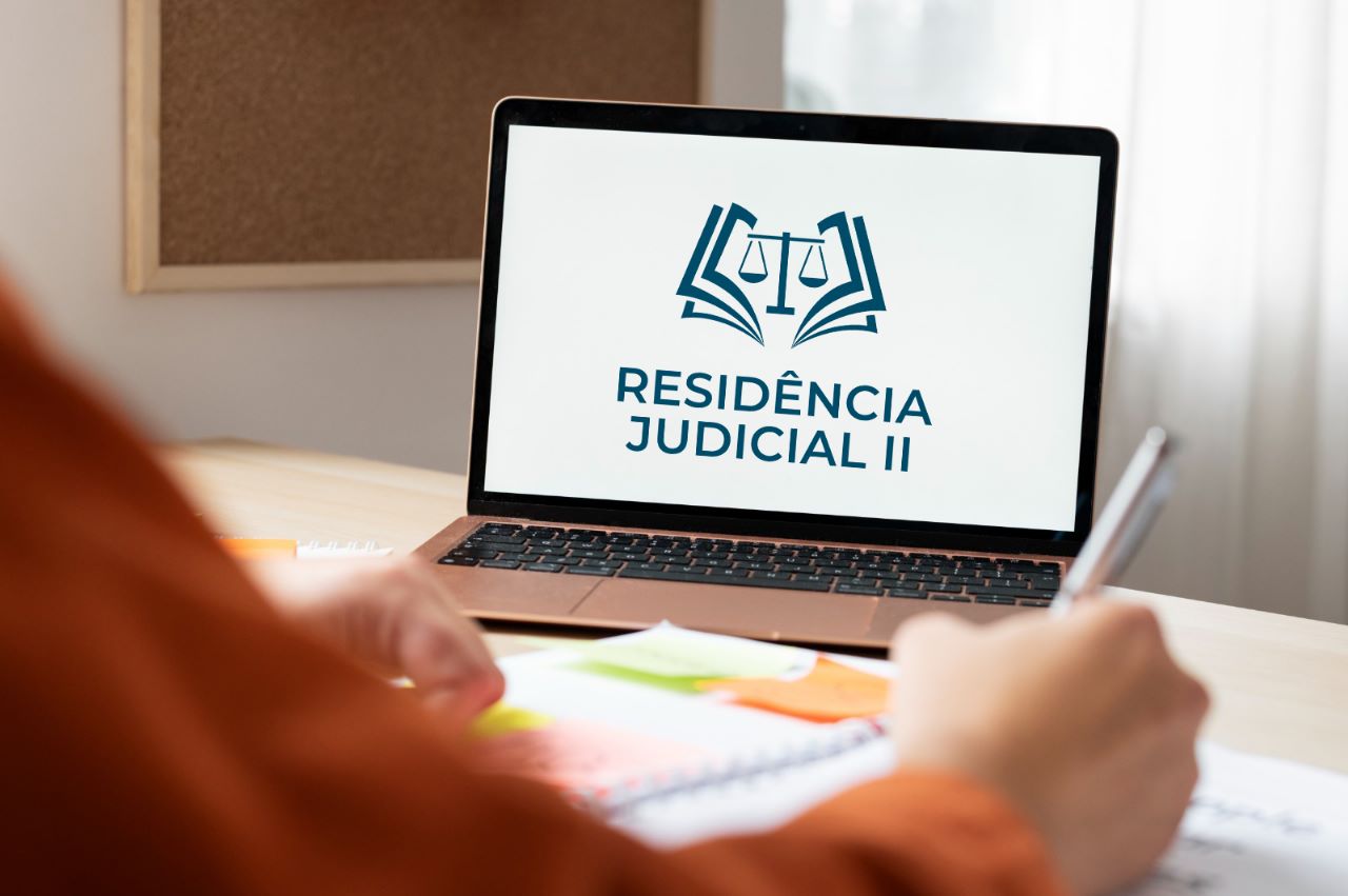 foto colorida mostra um notebook em cima de uma mesa. Na tela do aparelho está escrito "RESIDÊNCIA JUDICIAL” e acima a logomarca do programa. Em primeiro plano aparece de forma embaçada as mãos de uma pessoa escrevendo com uma caneta.