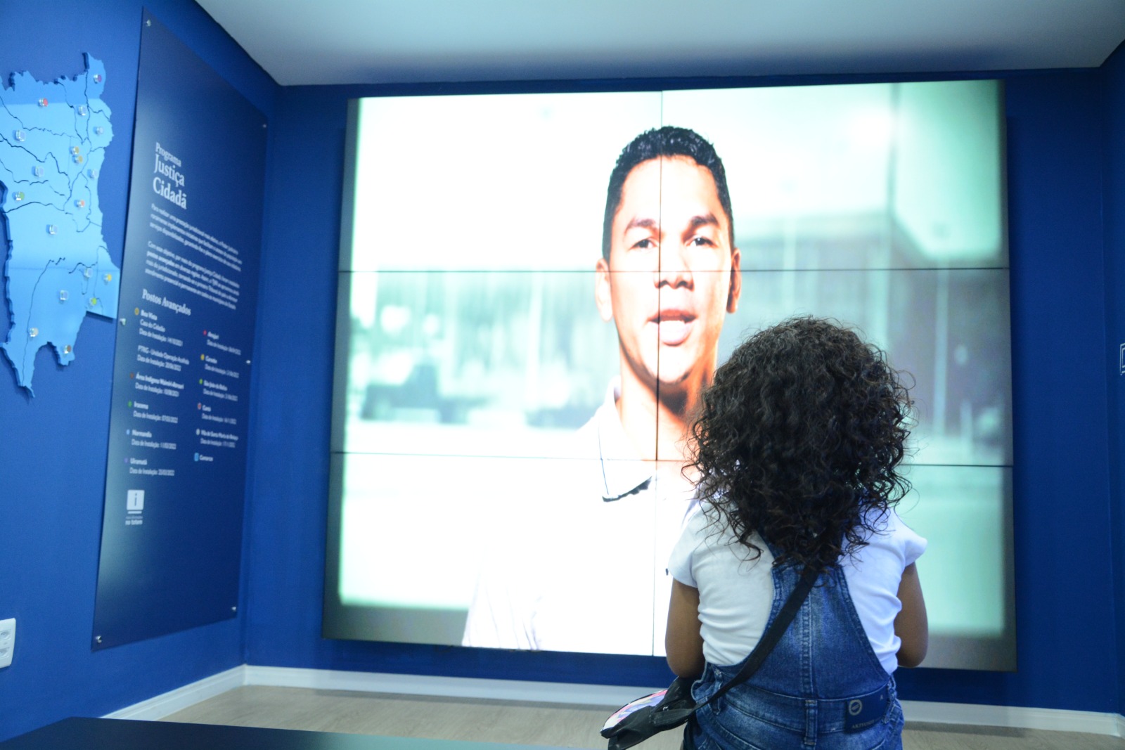 foto colorida mostra uma menina sentada de costas para a foto, observando um telão no Centro de Memória e Cultura do Poder Judiciário de Roraima. Na tela aparece o historiador do TJRR, Hugo Mendes, explicando a fundação do Brasil. 