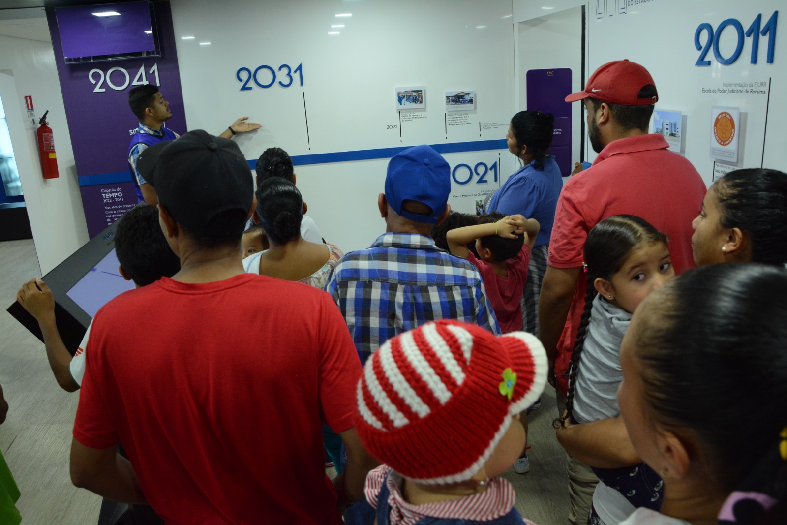  foto colorida mostra famílias de imigrantes venezuelanos, entre homens, mulheres e crianças, acompanhando a visita na linha do tempo do Poder Judiciário de Roraima. Um guia do Centro de Memória e Cultura do Poder Judiciário de Roraima faz a explicação. 