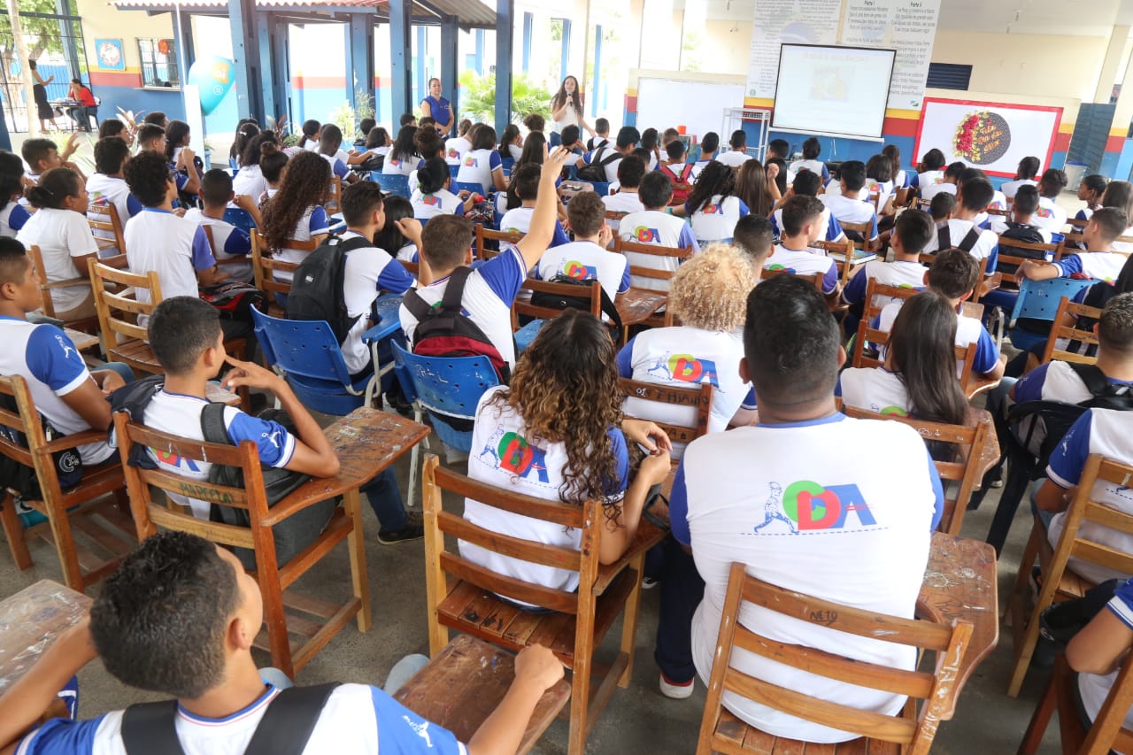  foto colorida mostra uma plateia de alunos, fardados, sentados em carteiras de madeiras, de costas para a foto, acompanhando a palestra ministrada pela coordenadora do programa Justiça Comunitária, Marcelle Wottrich, que está de pé a frente dos estudantes.