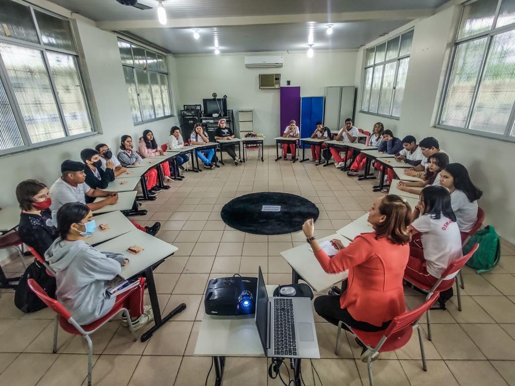 foto colorida mostra 20 alunos sentados frente a cadeiras organizadas em formato de círculo em uma sala de aula. Uma professora, também sentada no círculo, ministra palestra.