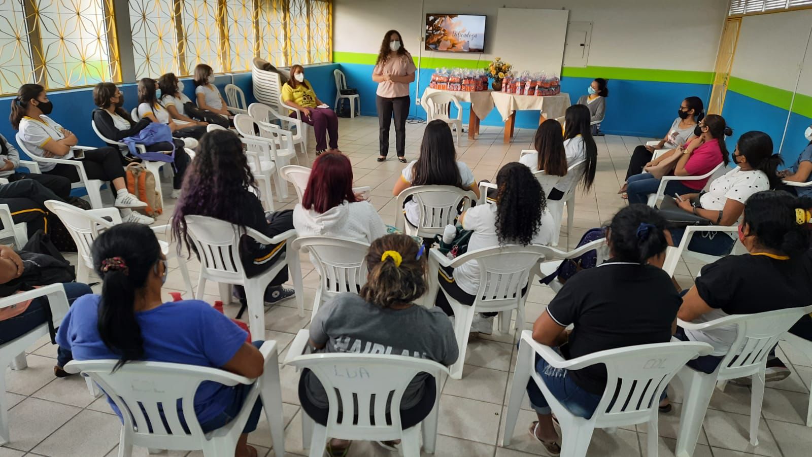  foto colorida mostra alunos sentados em cadeiras organizadas em formato de “c” acompanhando a palestra ministrada pela coordenadora do programa Justiça Comunitária, Marcelle Wottrich, que está de pé. Ao lado dela está uma mesa com panetones em cima. 