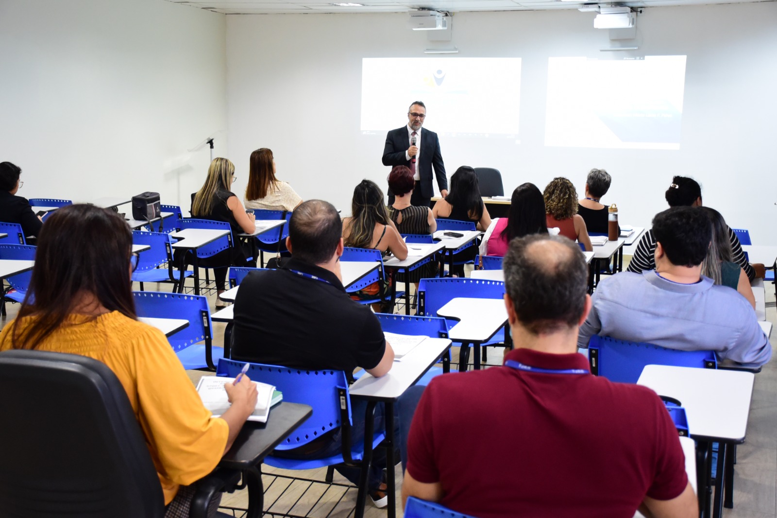 foto colorida mostra o instrutor da empresa TNQ - Consultoria e Treinamento, Mário Lúcio, em pé, falando ao microfone durante o treinamento. Cerca de 15 pessoas estão sentadas em cadeiras azuis, de costas para a foto, acompanhando a fala do instrutor.