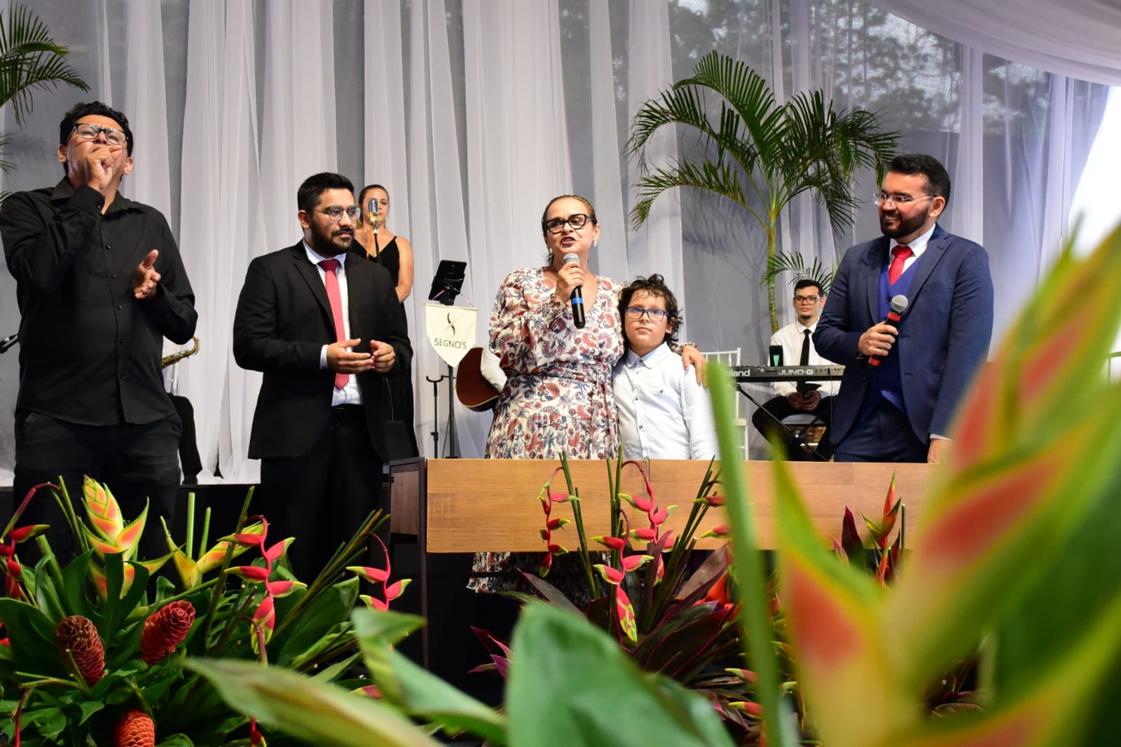 foto colorida mostra a juíza Graciete Sotto Mayor posicionada atrás de uma mesa, em pé, abraçando uma criança enquanto fala ao microfone durante a cerimônia de casamento coletivo. Ao lado dela estão os juízes de paz e o intérprete de libras.