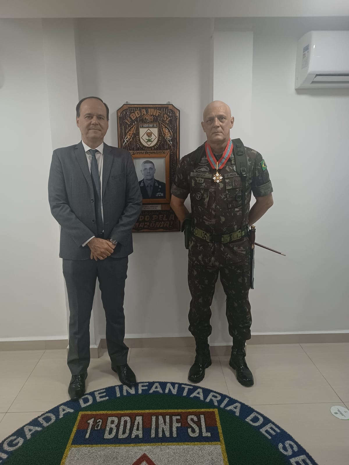 Imagem mostra o desembargador e o general de brigada Marcelo Lorenzini Zucco posando lado a lado. O desembargador está de terno e gravata em tons de cinza. Ele é um homem alto, de cabelos pretos, e posa com as mãos em frente ao corpo. Já o general está com farda do exército e uma medalha de honra no pescoço. Ele é um homem careca, e posa com as mãos para trás. Atrás deles, na parede, está um quadro com uma foto antiga e uma moldura de madeira. No chão, um tapete redondo escrito o nome da 1ª Brigada de Infantaria de Selva.