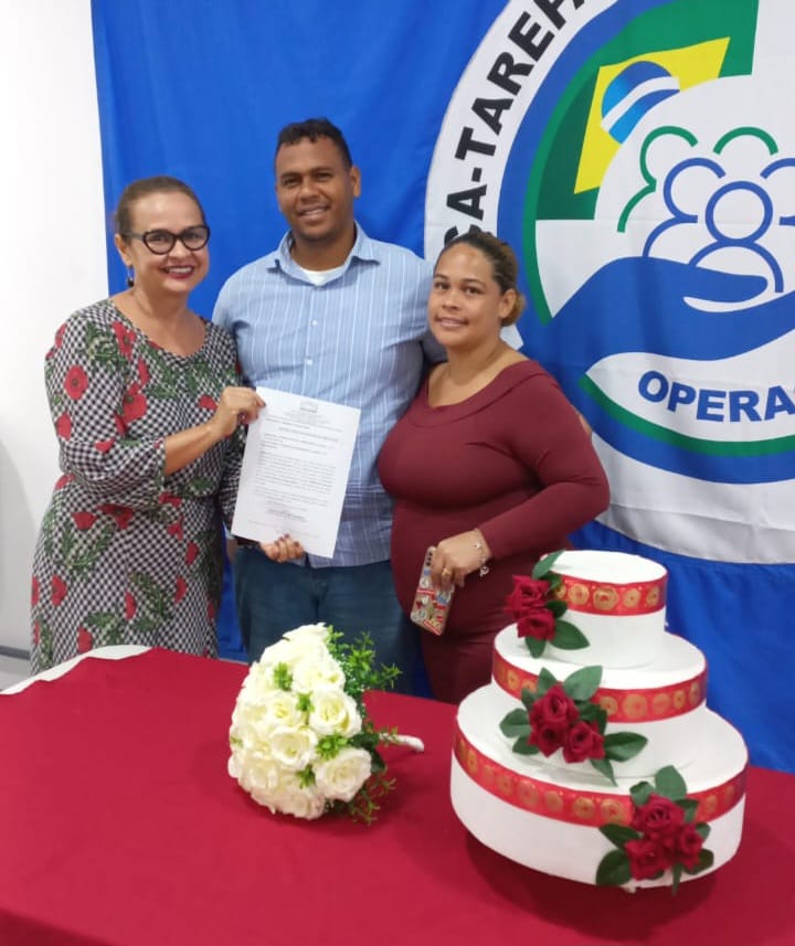 foto colorida mostra a juíza titular da Vara da Justiça Itinerante, Graciete Sotto Mayor, posando para foto ao lado de um casal e segurando a certidão de união estável dos mesmos. Todos estão atrás de uma mesa coberta por um tecido vermelho. Em cima do móvel está um bolo e um buquê de flores.