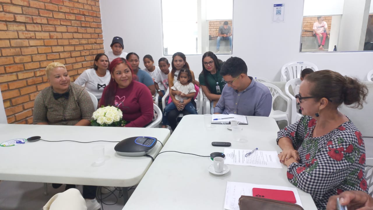 foto colorida mostra a direita a juíza titular da Vara da Justiça Itinerante, Graciete Sotto Mayor, sentada em frente a uma mesa enquanto realiza atendimentos juntamente a outro homem que está ao lado. Do lado esquerdo está um casal de mulheres sendo atendidas e outras nove pessoas, entre mulheres e crianças, aguardando o atendimento. 